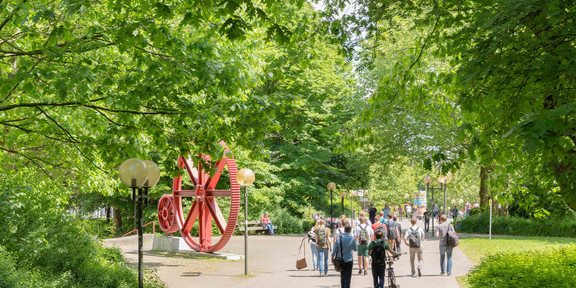 TU Dortmund im Sommer_04 Studierende laufen am Zahnrad vorbei und grüne Bäume säumen den Weg.