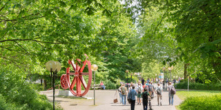 TU Dortmund im Sommer_04 Studierende laufen am Zahnrad vorbei und grüne Bäume säumen den Weg.