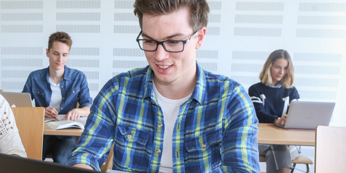 Student sitzt im Seminarraum am Laptop, im Hintergrund weitere Studierende